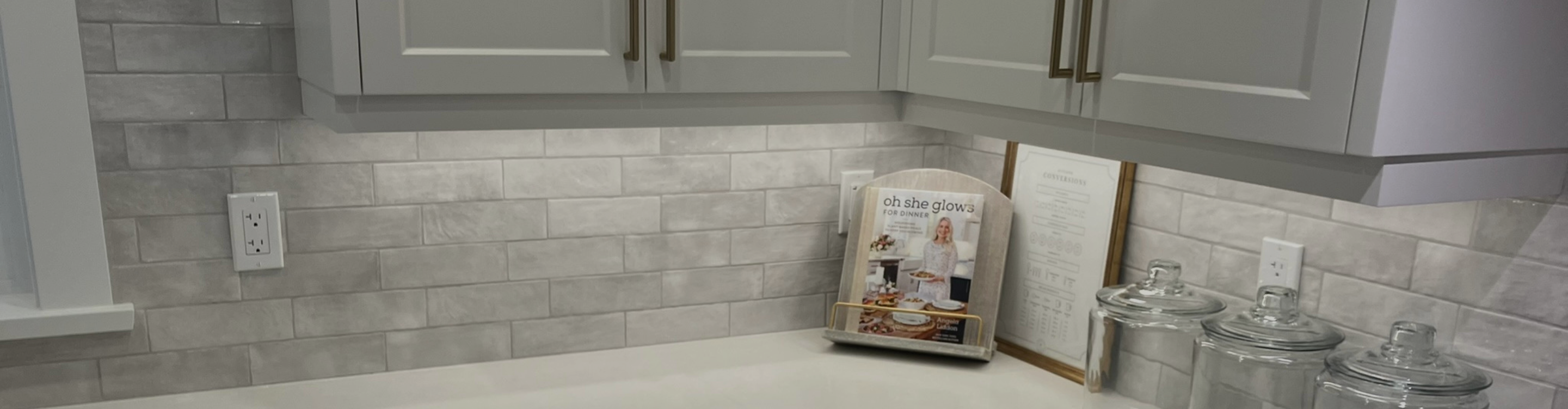 A kitchen counter with white quartz countertops, light gray cabinets with brushed gold hardware, and a subway tile backsplash. A cookbook titled "Oh She Glows" is propped open on the counter.